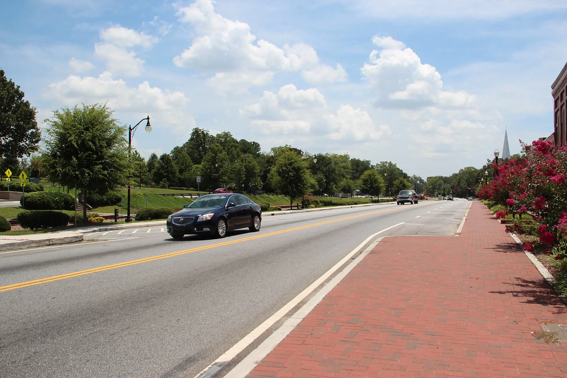 Main Street in downtown Jonesboro on a summer day: a two-lane road with a brick sidewalk, crape myrtles in bloom and a church steeple in the distance